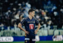 Frühe Führung reicht nicht: FCL unterliegt Meister Thun Soccer player in a blue and gold kit stands with hands on hips on a grassy pitch, blurred stadium crowds behind him.