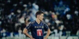 Soccer player in a blue and gold kit stands with hands on hips on a grassy pitch, blurred stadium crowds behind him.