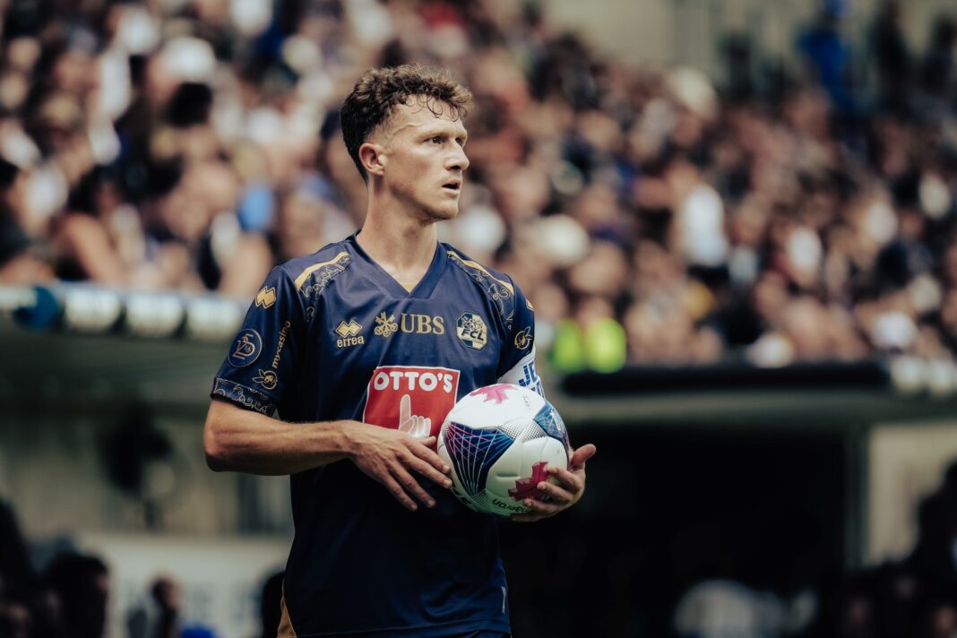 dsc019893 Soccer player in a blue jersey holding a white ball, stadium crowd blurred in the background.