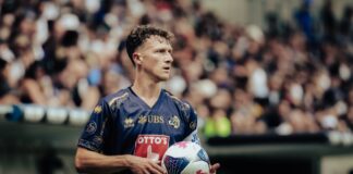 Soccer player in a blue jersey holding a white ball, stadium crowd blurred in the background.