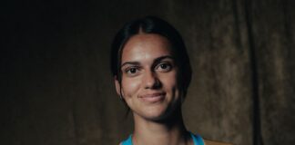 Portrait of a smiling woman with dark hair in a blue athletic jersey, against a dark textured background.