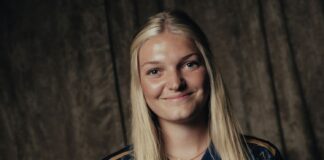 Smiling young woman with long blonde hair wearing a navy sports jersey, standing against a dark textured backdrop.