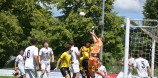 Soccer game near the goal with an orange-clad goalkeeper jumping to punch the ball away amid white and yellow players nearby.