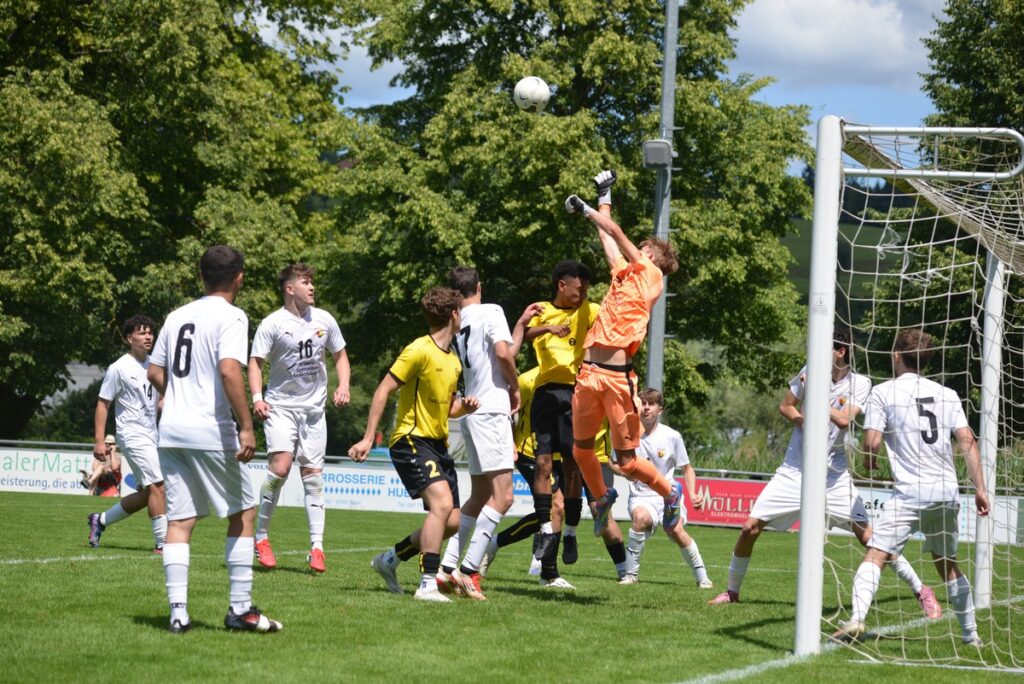 fcbaar_svmeggenadligenswil18-1024x684 Soccer game near the goal with an orange-clad goalkeeper jumping to punch the ball away amid white and yellow players nearby.