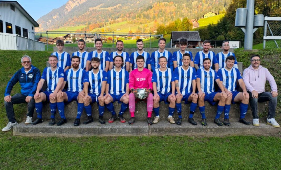 fclungenr Group of soccer players in blue-and-white striped uniforms posing on concrete steps with a pink-clad goalkeeper holding a ball; coaches flank them, rural field backdrop behind.