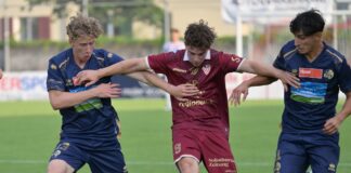 Three soccer players in blue and maroon kits compete for the ball on a green field during a match, with the central maroon player shielding the ball from two opponents.