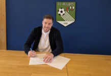 Joel Stephan neuer Spielertrainer beim FC Algro Man sits at a wooden table signing a document with a blue wall and a green FC ALGRO crest in the background.