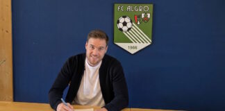 Man sits at a wooden table signing a document with a blue wall and a green FC ALGRO crest in the background.