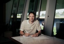 FC Luzern: Levin Winkler verlängert vorzeitig bis 2029 Man sits at a marble table signing a document with a pen in a bright, modern room with large windows behind him and daylight coming in.