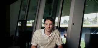 Man sits at a marble table signing a document with a pen in a bright, modern room with large windows behind him and daylight coming in.