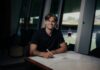 FC Luzern verpflichtet Loïc Lüthi Young man with tattoos smiling while signing documents at a marble table in a modern glass-walled room, with a sports stadium visible through the windows outside.