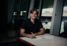 FC Luzern verpflichtet Loïc Lüthi Young man with tattoos smiling while signing documents at a marble table in a modern glass-walled room, with a sports stadium visible through the windows outside.