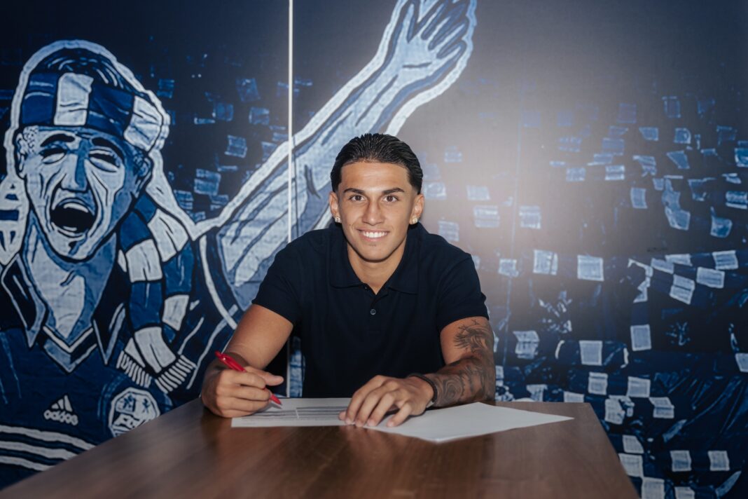 Young man smiles while signing a document at a table, blue mural backdrop behind him