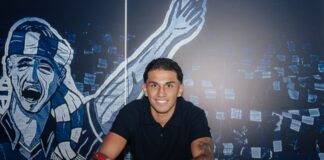 Young man smiles while signing a document at a table, blue mural backdrop behind him