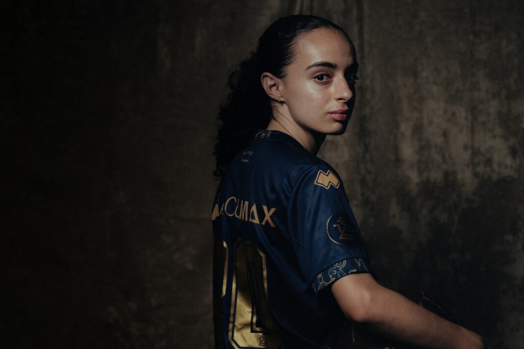 michelle-gigante Portrait of a person in a blue and gold sports jersey, turned to look over their shoulder against a dark textured background.
