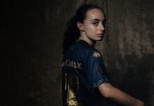 FCL: Michelle Gigante stösst zum AWSL-Team Portrait of a person in a blue and gold sports jersey, turned to look over their shoulder against a dark textured background.
