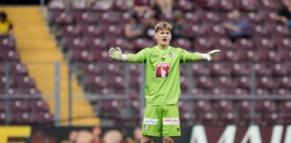 Soccer goalkeeper in neon green kit stands with arms outstretched on the pitch during a match.