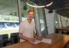 Rückkehrer: FC Luzern verpflichtet Raphael Radtke Smiling man signs a document at a wooden table in a glass-walled office, with stadium seating visible outside.