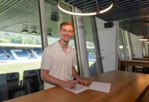 Rückkehrer: FC Luzern verpflichtet Raphael Radtke Smiling man signs a document at a wooden table in a glass-walled office, with stadium seating visible outside.