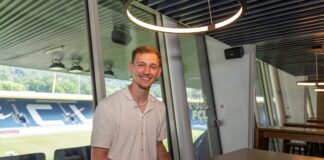 Smiling man signs a document at a wooden table in a glass-walled office, with stadium seating visible outside.