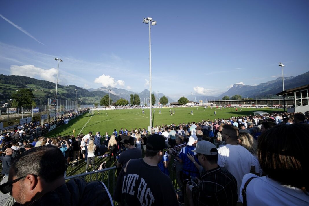 Crowd watching a daytime soccer match at a sunny stadium with green field and mountain backdrop.