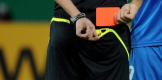 Referee in black shorts with neon trim holding an orange card during a soccer match, with a blue-player nearby.