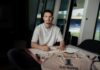 FC Luzern verpflichtet Simon Simoni Man in a white T-shirt signs documents at a table, with a sports jersey spread out in front and stadium seating visible through the windows behind him.