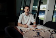 FC Luzern verpflichtet Simon Simoni Man in a white T-shirt signs documents at a table, with a sports jersey spread out in front and stadium seating visible through the windows behind him.