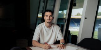 Man in a white T-shirt signs documents at a table, with a sports jersey spread out in front and stadium seating visible through the windows behind him.