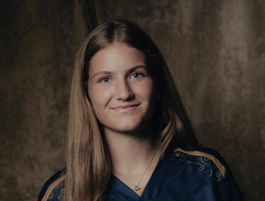 valentina-gerlof-awsl-team Portrait of a young woman with long brown hair smiling at the camera, wearing a navy top with a pendant necklace, against a dark studio backdrop.