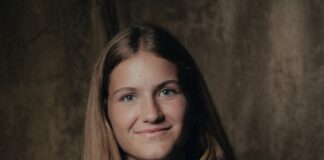 Portrait of a young woman with long brown hair smiling at the camera, wearing a navy top with a pendant necklace, against a dark studio backdrop.