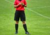 IFV kritisiert FIFA scharf: Fussball darf nicht politisch beeinflusst werden Soccer referee on a green field in a red jersey and black shorts, holding a yellow card and a notebook.