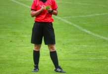 IFV kritisiert FIFA scharf: Fussball darf nicht politisch beeinflusst werden Soccer referee on a green field in a red jersey and black shorts, holding a yellow card and a notebook.
