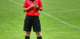 Soccer referee on a green field in a red jersey and black shorts, holding a yellow card and a notebook.
