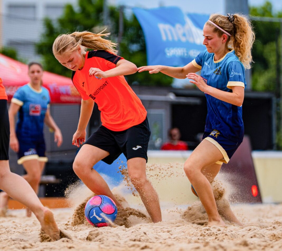 Two women play beach soccer on a sandy court, one in orange and black and the other in blue, chasing a colorful ball.