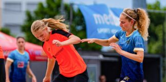 Two women play beach soccer on a sandy court, one in orange and black and the other in blue, chasing a colorful ball.