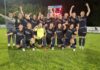 TRE-Frauen lassen sich den Sieg nicht nehmen Women’s soccer team posing for a celebration photo on a grass field at night, navy uniforms with sponsor logos, goalkeeper in neon yellow front center, players raising arms in victory.