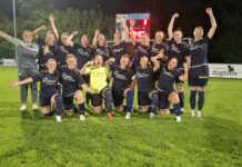 TRE-Frauen lassen sich den Sieg nicht nehmen Women’s soccer team posing for a celebration photo on a grass field at night, navy uniforms with sponsor logos, goalkeeper in neon yellow front center, players raising arms in victory.