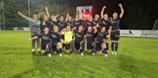 Women’s soccer team posing for a celebration photo on a grass field at night, navy uniforms with sponsor logos, goalkeeper in neon yellow front center, players raising arms in victory.