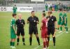 Die grosse Vorschau: Perlen-Buchrain startet in die neue Saison Referees in black uniforms converse with a maroon-clad player on a green soccer field, with teammates in green waiting nearby on the sideline.