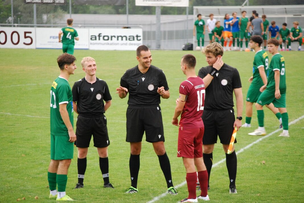 Referees in black uniforms converse with a maroon-clad player on a green soccer field, with teammates in green waiting nearby on the sideline.