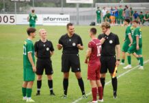 Die grosse Vorschau: Perlen-Buchrain startet in die neue Saison Referees in black uniforms converse with a maroon-clad player on a green soccer field, with teammates in green waiting nearby on the sideline.