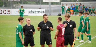 Referees in black uniforms converse with a maroon-clad player on a green soccer field, with teammates in green waiting nearby on the sideline.