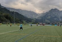 Engelberg überrennt Adligenswil Soccer match on a green field with players in yellow and teal jerseys, mountains in the background under cloudy skies.