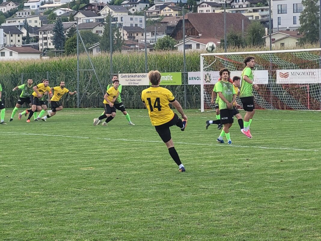 Soccer player in a yellow jersey (#14) kicks the ball toward the goal while teammates and green-clad players react on a grassy field.