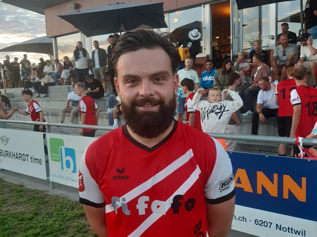 Bearded man in a red and white diagonal-striped sports jersey stands in front of spectators at an outdoor stadium.