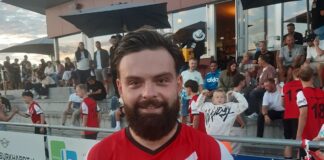 Bearded man in a red and white diagonal-striped sports jersey stands in front of spectators at an outdoor stadium.