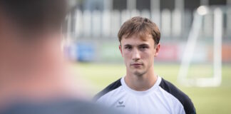 Portrait of a young male athlete in a white and black jersey, facing the camera on a grassy soccer field with a blurred teammate in the foreground.