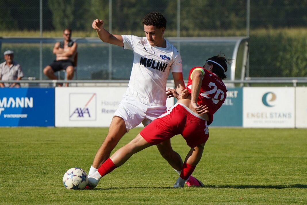 3 Soccer players in white and red compete for the ball on a sunny field, white jersey shielding a low kick from a red-jersey opponent.