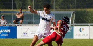 Soccer players in white and red compete for the ball on a sunny field, white jersey shielding a low kick from a red-jersey opponent.